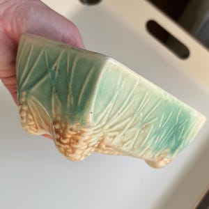 以下が含まれることがあります： 明るい緑と茶色の小さな長方形のセラミックプランター。プランターは葉と松ぼっくりのテクスチャデザインが特徴です。3つの小さな足があります。