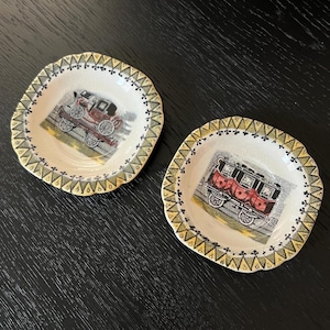 May include: Two small, decorative ceramic dishes with scalloped edges. Each dish features a vintage illustration of a horse-drawn carriage in red and black. The rims are decorated with a repeating pattern in yellow and green.
