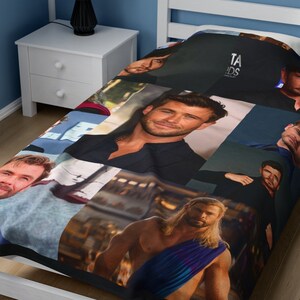 May include: A black and blue fleece blanket with a collage of images of a man with long blonde hair. The images are arranged in a grid pattern and include close-ups of his face, as well as full-body shots. The blanket is folded and displayed on a white bed frame.
