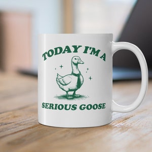 Può includere: Una tazza in ceramica bianca con un'illustrazione verde di un'oca con una cravatta. Il testo "TODAY I'M A SERIOUS GOOSE" è stampato sulla tazza.