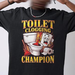 May include: Black t-shirt with a humorous graphic. The design features a toilet, plunger, and animated toilet paper roll with the text "TOILET CLOGGING CHAMPION" in yellow and orange.