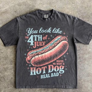 May include: Dark gray t-shirt with a graphic of a hot dog and the text "You look like the 4th of July makes me want a hot dog real bad." The design features red, blue, and white colors.