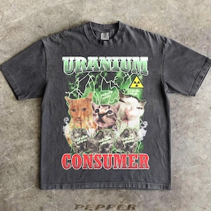 May include: Dark gray t-shirt with a graphic featuring three cats and the words "URANIUM CONSUMER" in green and red. The design includes a radiation symbol and a lightning bolt pattern.