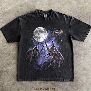 Op de afbeelding: Een zwart t-shirt met korte mouwen en een grafische print van drie neushoornkevers tegen een achtergrond van een volle maan en een paars-blauw sterrenstelsel. Het shirt heeft een ronde hals en een vervaagde look over de hele stof.
