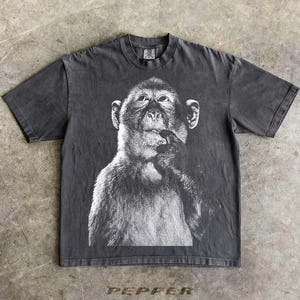以下が含まれることがあります： 黒と白の猿の画像がプリントされた濃い灰色のTシャツ。猿は思慮深いポーズで描かれており、手は口の近くにあります。「PEPPER」の文字がシャツの下部に印刷されています。