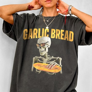 Könnte beinhalten: Ein schwarzes Oversize-T-Shirt mit einem Aufdruck eines Skeletts, das eine Sonnenbrille trägt und ein Knoblauchbrot hält. Der Text "GARLIC BREAD" ist in Gold über dem Skelett gedruckt.