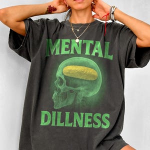 May include: A black t-shirt featuring a green skull graphic with the text "MENTAL DILLNESS". The skull has a yellow pickle inside of it.