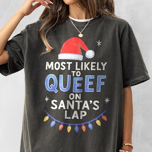 May include: Dark gray t-shirt with a Christmas-themed graphic. The design includes a red Santa hat, snowflakes, and the text "MOST LIKELY TO QUEEF ON SANTA'S LAP" in blue and white. A string of colorful lights is at the bottom.