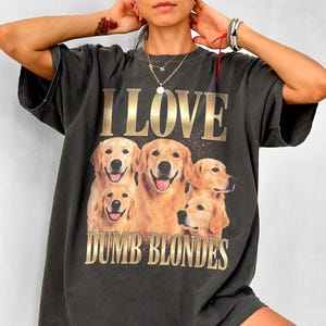 May include: Dark gray t-shirt featuring a graphic of five golden retriever dogs with the text "I LOVE DUMB BLONDES" in gold lettering. The shirt has short sleeves and a relaxed fit, suitable for casual wear.