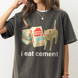 May include: A dark gray oversized t-shirt with a graphic print of a cat eating bags of cement. The text "i eat cement" is printed below the image.
