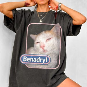 Może przedstawiać: Ciemnoszary t-shirt z krótkim rękawem z nadrukiem graficznym przedstawiającym białego i rudego kota z przymrużonymi oczami w prostokącie z różową obwódką. Pod kotem znajduje się niebieska owalna odznaka z białym napisem Benadryl pogrubioną czcionką.