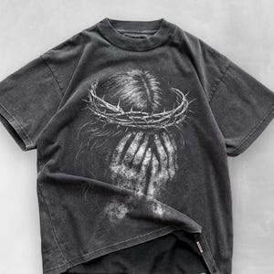 May include: Dark gray t-shirt featuring a distressed graphic of a person with a crown of thorns and hands covering their face. The design is in white, creating a striking contrast against the shirt's dark background. The shirt has short sleeves and a crew neck.