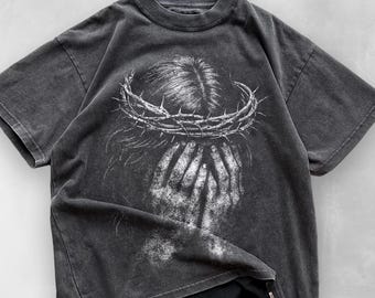 May include: Dark gray t-shirt featuring a distressed graphic of a person with a crown of thorns and hands covering their face. The design is in white, creating a striking contrast against the shirt's dark background. The shirt has short sleeves and a crew neck.