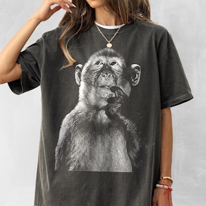 Peut inclure: T-shirt gris foncé avec une illustration en noir et blanc d'un singe. Le singe est représenté dans une pose pensive, la main près de la bouche. Le t-shirt a des manches courtes et une coupe ample, adapté pour un usage décontracté.