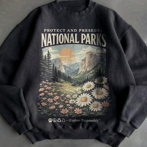 May include: Dark grey sweatshirt featuring a mountain landscape graphic with flowers and the text "PROTECT AND PRESERVE NATIONAL PARKS". The design includes a sunset, trees, and "Explore Responsibly".