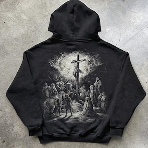May include: Black hooded sweatshirt featuring a detailed grayscale illustration on the back depicting a crucifixion scene. The artwork includes figures, a cross, and a dramatic sky. The word "BLACK" is printed at the bottom.