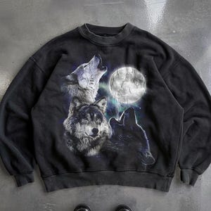 May include: Black sweatshirt featuring a graphic of three wolves howling at a full moon. The wolves are depicted in shades of gray and white, with the moon in the background. The sweatshirt has a crew neck and long sleeves.