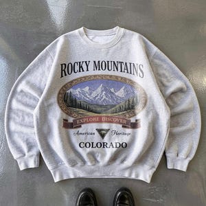 Op de afbeelding: Een grijze crewneck sweatshirt met een afbeelding van de Rocky Mountains. Het ontwerp bevat een ovaal beeld van bergen en bomen, met de tekst "ROCKY MOUNTAINS", "EXPLORE DISCOVER", "American Heritage" en "COLORADO".