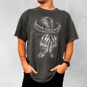 May include: Dark gray t-shirt featuring a distressed graphic of a crown of thorns and hands covering a face. The design is in white, creating a high-contrast effect. The shirt has a vintage, worn look.