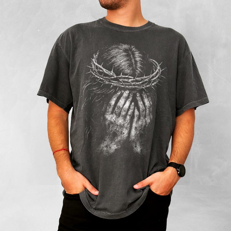 May include: Dark gray t-shirt featuring a distressed graphic of a crown of thorns and hands covering a face. The design is in white, creating a high-contrast effect. The shirt has a vintage, worn look.