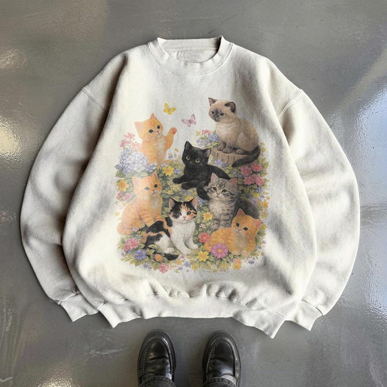 May include: Cream-colored sweatshirt featuring a colorful illustration of several kittens surrounded by flowers and butterflies. The kittens are in various poses and colors, including orange, black, and calico. The sweatshirt has a crew neck and long sleeves.
