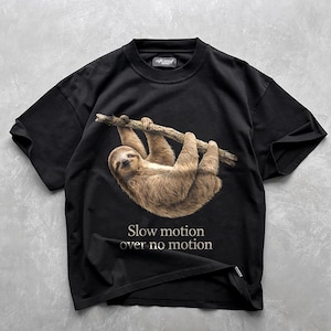 Peut inclure: Un t-shirt noir à manches courtes orné d'un graphisme réaliste représentant un paresseux brun suspendu à une branche d'arbre. Sous le paresseux, le texte "Slow motion over no motion" est imprimé dans une police serif claire. Col rond.