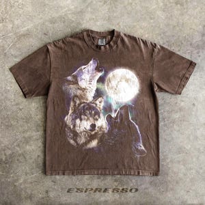 K&ouml;nnte beinhalten: Braunes T-Shirt mit einer Grafik von drei W&ouml;lfen, die einen Vollmond anheulen. Das Design beinhaltet Lila- und Wei&szlig;t&ouml;ne, mit dem Wort "ESPRESSO" unten aufgedruckt.