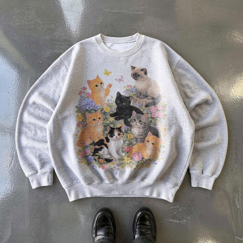 May include: A light gray sweatshirt featuring a colorful graphic of several cats surrounded by flowers and butterflies. The cats are in various colors, including orange, black, and gray. The sweatshirt has a crew neck and long sleeves.