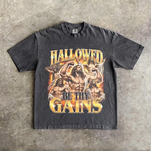 Könnte beinhalten: Ein anthrazitfarbenes Kurzarm-T-Shirt mit einem Grafikdruck von muskulösen Figuren mit langem Haar und Bärten vor einem Hintergrund aus orangefarbenen Flammen. Der Text HALLOWED BE THY GAINS ist in großen, goldenen Buchstaben gedruckt.