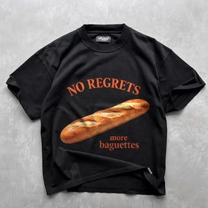 May include: A black short-sleeve t-shirt featuring a realistic image of a golden-brown baguette centered on the chest. Above the bread, the text reads NO REGRETS in orange, and below the bread, the text reads more baguettes in orange lettering.