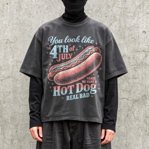 May include: Dark gray t-shirt with a vintage-style hot dog graphic. The text on the shirt reads "You look like the 4th of July makes me want a hot dog real bad."