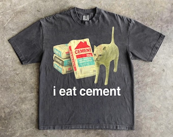 May include: Dark gray t-shirt with a graphic featuring a stack of cement bags, a cat, and the text "i eat cement". The cement bags are beige with red and blue details. The cat is light green.