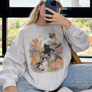 May include: A light gray sweatshirt featuring a colorful graphic of several kittens surrounded by flowers and butterflies. The kittens are in various poses and colors, including orange, black, and gray. The sweatshirt has long sleeves.