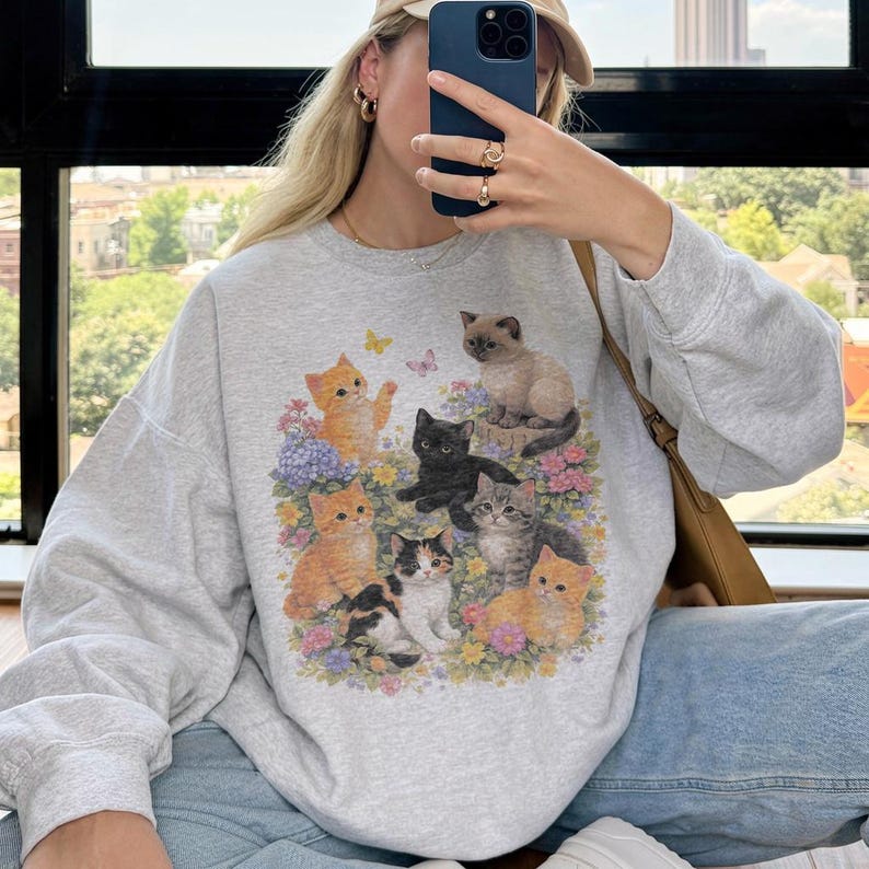 May include: A light gray sweatshirt featuring a colorful graphic of several kittens surrounded by flowers and butterflies. The kittens are in various poses and colors, including orange, black, and gray. The sweatshirt has long sleeves.