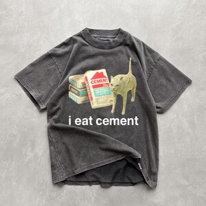 May include: A black t-shirt with a distressed look features a white cat looking at a stack of cement bags. The text "i eat cement" is printed below the cat.