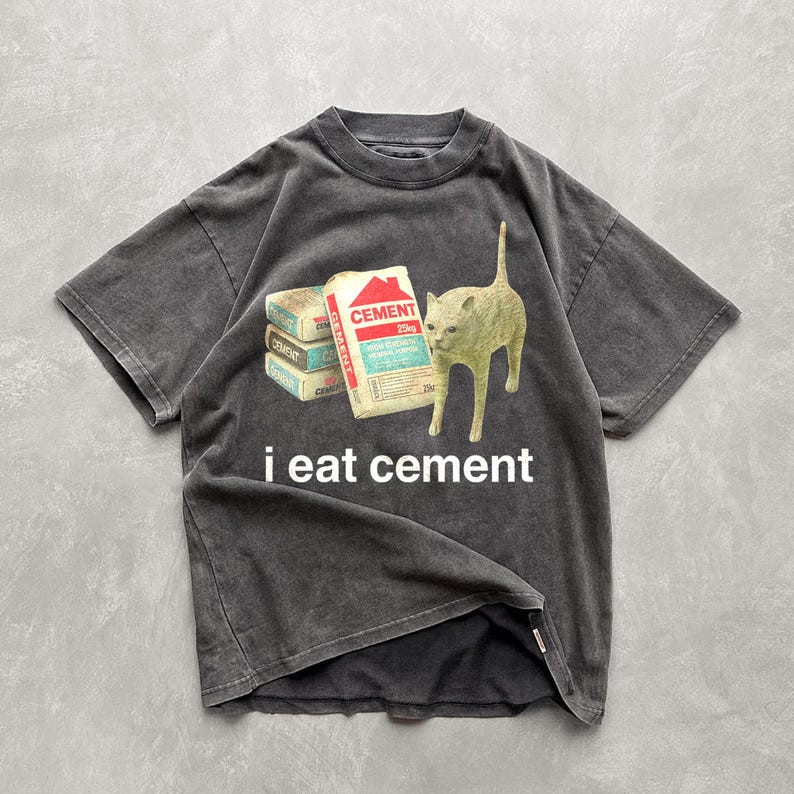 May include: A black t-shirt with a distressed look features a white cat looking at a stack of cement bags. The text "i eat cement" is printed below the cat.