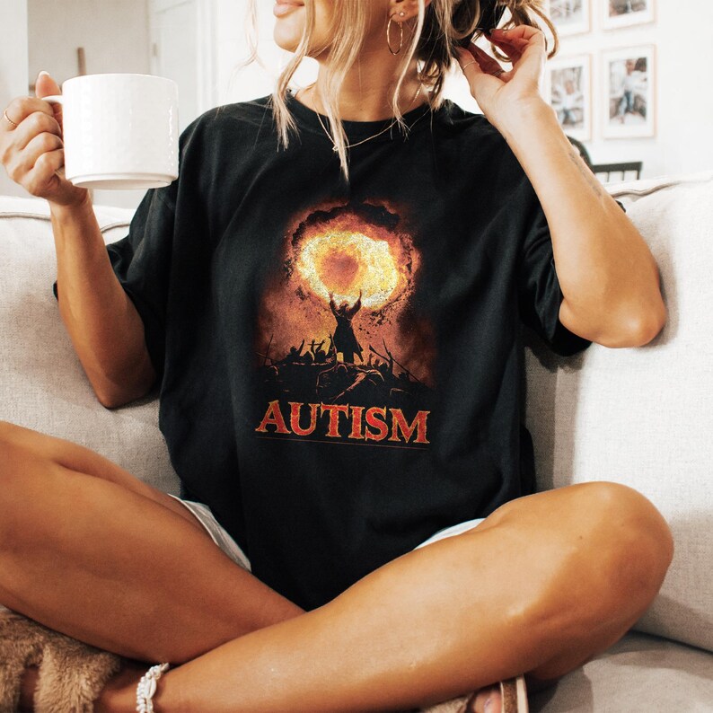 Puede incluir: Camiseta negra con un gr&aacute;fico de una persona sosteniendo un c&iacute;rculo en llamas sobre su cabeza. El texto "AUTISM" est&aacute; impreso debajo del gr&aacute;fico.