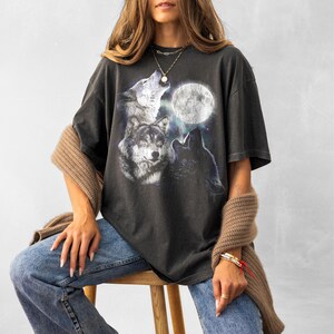 K&ouml;nnte beinhalten: Dunkelgraues T-Shirt mit einer Grafik von drei W&ouml;lfen, die den Vollmond anheulen. Das Design umfasst Graut&ouml;ne, Wei&szlig; und Blau. Das Shirt wird mit einer blauen Jeans und einem braunen Pullover getragen.