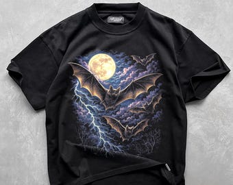 May include: A black short-sleeve t-shirt featuring a graphic print of three bats flying through a night sky with a full moon, dark purple clouds, and a bright lightning bolt striking down toward silhouettes of bare trees at the bottom.