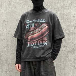 May include: Dark gray t-shirt with a distressed graphic of a hot dog and the text "You look like the 4th of July makes me want a hot dog real bad." The design features red, blue, and white colors.