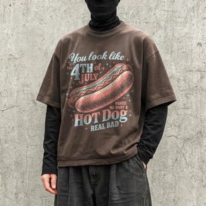 May include: Brown t-shirt with a distressed graphic featuring a hot dog and the text "You look like the 4th of July makes me want a hot dog real bad." The shirt has short sleeves.