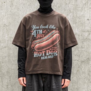 May include: Brown t-shirt with a graphic of a hot dog and the text "You look like the 4th of July makes me want a hot dog real bad". The shirt has short sleeves and a relaxed fit.