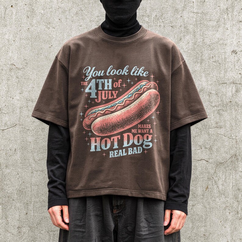 May include: Brown t-shirt with a graphic of a hot dog and the text "You look like the 4th of July makes me want a hot dog real bad". The shirt has short sleeves and a relaxed fit.