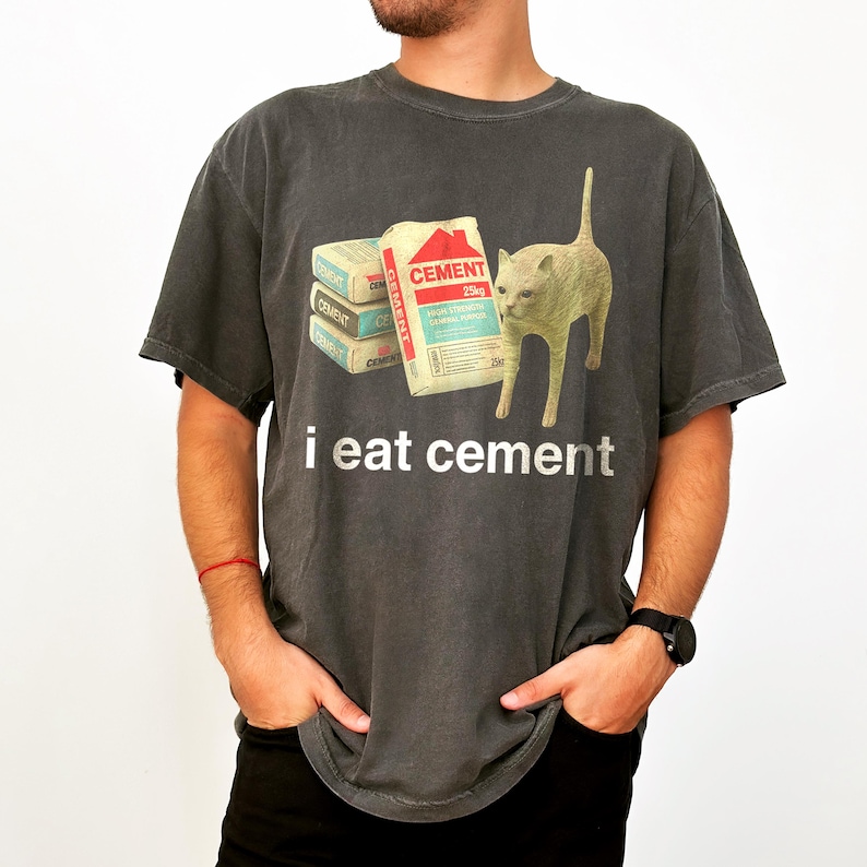 May include: A dark gray t-shirt with a graphic of a cat standing next to a stack of cement bags. The text "i eat cement" is printed below the graphic.