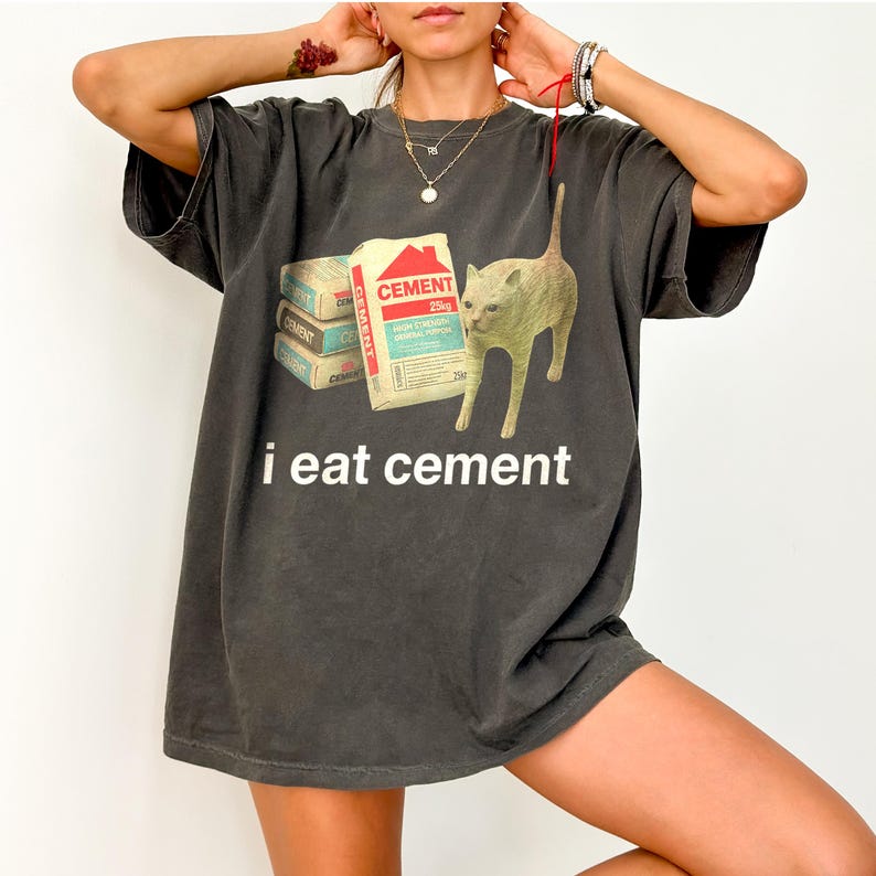 May include: A dark gray oversized t-shirt with a graphic of a cat looking at a stack of cement bags. The text "i eat cement" is printed below the graphic.