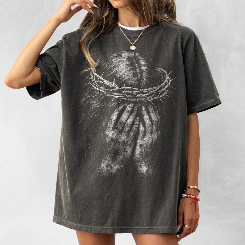 May include: Dark gray t-shirt featuring a distressed graphic of a crown of thorns above a pair of hands. The design is in white, creating a stark contrast against the shirt's color. The shirt has a classic crew neck and short sleeves, offering a relaxed fit.