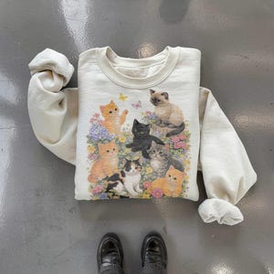 May include: Cream-colored sweatshirt featuring a colorful graphic of several cats amidst flowers and butterflies. The cats are in various poses and colors, including orange, black, and gray. The sweatshirt has a crew neck and long sleeves.