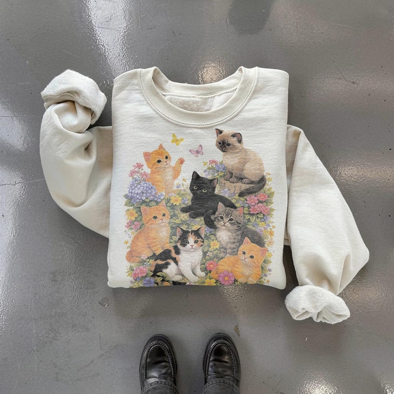 May include: Cream-colored sweatshirt featuring a colorful graphic of several cats amidst flowers and butterflies. The cats are in various poses and colors, including orange, black, and gray. The sweatshirt has a crew neck and long sleeves.