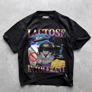 Può includere: T-shirt nero con un design grafico raffigurante un gatto con un elmetto, una bottiglia di latte con un segnale di divieto e una fetta di formaggio. Le parole "LACTOSE INTOLERANT" sono stampate sopra e sotto in un font viola e oro.