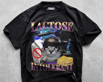 May include: Black t-shirt with a graphic design featuring a cat wearing a helmet, a bottle of milk with a no-entry sign, and a wedge of cheese. The words "LACTOSE INTOLERANT" are printed across the top and bottom in a purple and gold font.